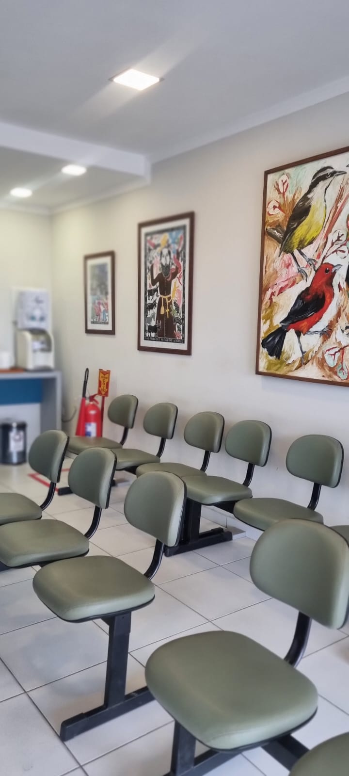 Sala de espera com cadeiras estofadas e quadros de arte regional pernambucana